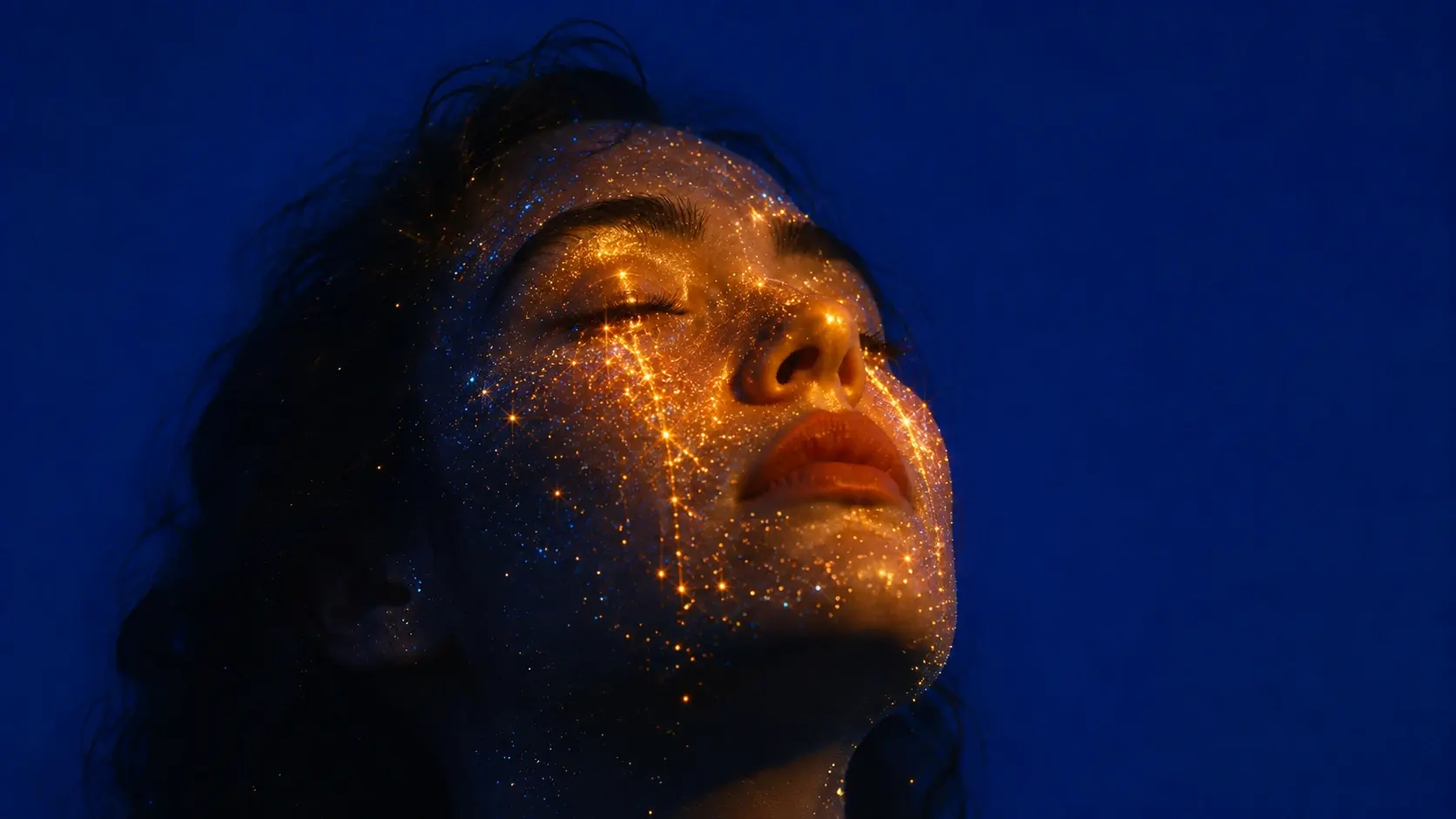 A close-up surreal portrait of a human face against a completely uninterrupted deep cobalt-blue background, the face turned slightly upward with eyes closed, several delicate streams of glowing golden particles flowing downward from the closed eyes like tears made from stardust, thousands of tiny blue-white and amber points of light scattered subtly across the cheeks and forehead, individual particles creating occasional tiny starburst flares, warm golden light illuminating only the cheekbones, nose and lips while most of the face remains surrounded by deep blue shadow, realistic skin and natural facial texture softened through a dreamy optical lens, emotional but serene expression, magical realism, intimate contemporary photography, cinematic contrast, subtle underexposure, warm amber against saturated cobalt blue, fine analog grain, gentle halation, slight chromatic softness, imperfect vintage lens rendering, atmospheric dust, poetic visual storytelling, restrained surrealism, no jewelry, no text, no typography, no graphic effects. COMPOSITION: face occupying the lower-middle portion of the frame, eyes angled upward.