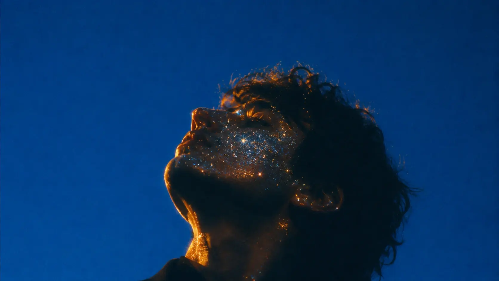 A close cinematic portrait of a human face in profile, head tilted upward toward an unseen source of light, the face partially illuminated by warm golden-orange rim light while the rest of the figure dissolves into deep cobalt-blue shadow, thousands of tiny luminous particles embedded naturally across the cheekbones, forehead, nose and jaw like an extremely subtle celestial constellation, individual particles sparkling with delicate white, blue and amber highlights, a handful of stronger star-shaped glints appearing across the skin, dark textured hair blending softly into the blue background, no visible studio environment, only an immense uninterrupted cobalt-blue atmosphere behind the subject, expression peaceful and contemplative, surreal magical realism, intimate photographic realism, rich skin texture softened by atmospheric diffusion, strong warm-versus-cool color contrast, subtle lens bloom around every bright particle, vintage optical character, restrained grain, faint chromatic aberration at high-contrast edges, slight underexposure in the shadows, cinematic tonal depth, poetic and mystical rather than glamorous, sophisticated contemporary editorial photography, no text, no typography, no decorative graphics. COMPOSITION: close-up profile occupying the lower-right portion of the frame.